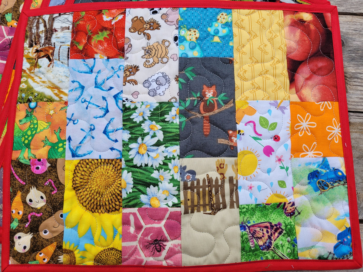 A square quilted fabric mat featuring a colorful patchwork design of various animal and nature prints, bordered by a red stitched edge, shown on a wooden surface.