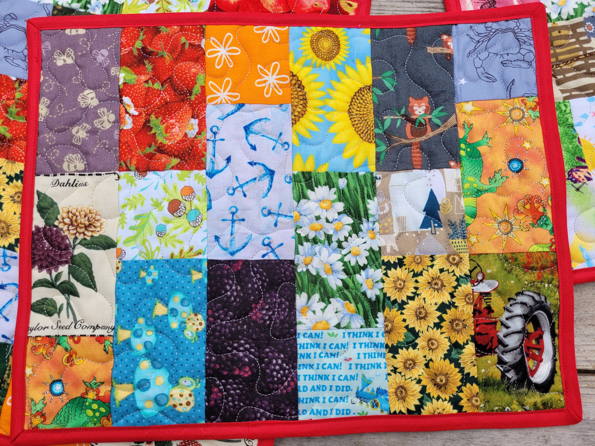 A square patchwork quilted fabric placemat, featuring a grid of colorful printed fabric squares with floral, sunflower, anchor, strawberry, and tractor motifs, finished with a red border.