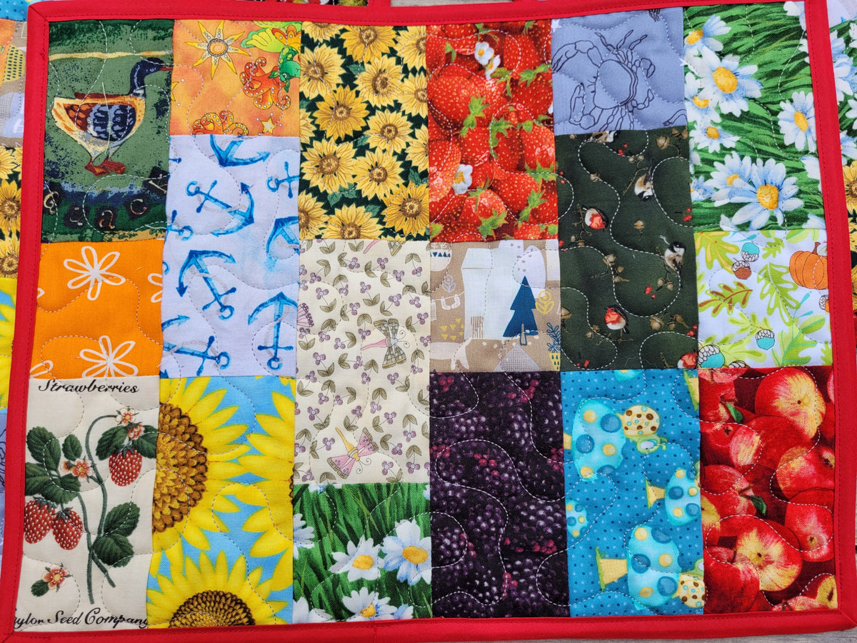 A square quilted fabric mat featuring a colorful patchwork grid of various fruit and botanical prints, finished with a solid red border. The patchwork includes designs of strawberries, sunflowers, raspberries, apples, daisies, and blue anchors.