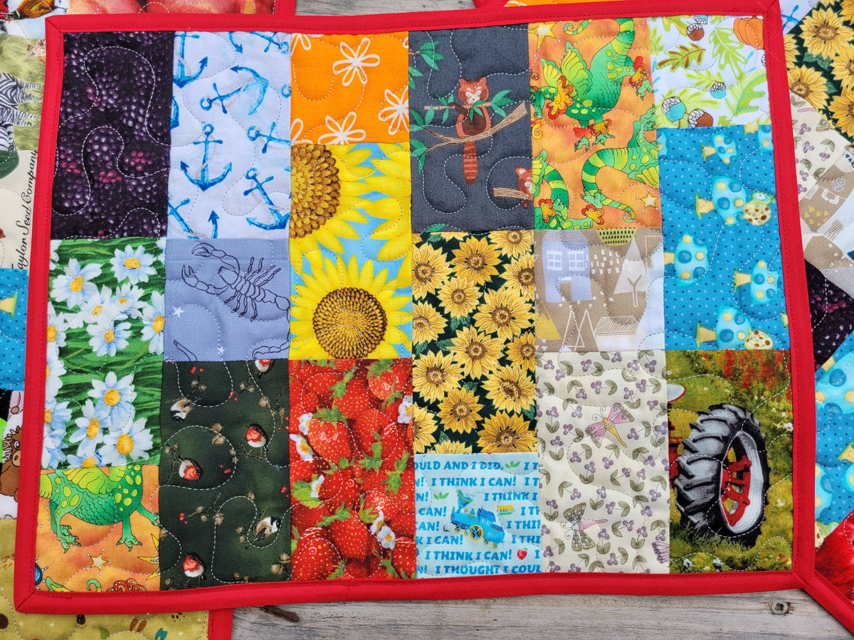 A square patchwork placemat with a red border, featuring various colorful fabric panels with patterns like sunflowers, strawberries, anchors, trees, and text prints, laid flat on a wooden surface.