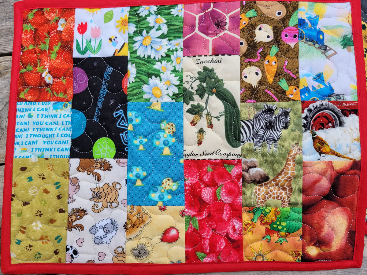 A quilted fabric I Spy placemat featuring a colorful patchwork design with various animal and nature-themed prints, finished with a solid red stitched border.