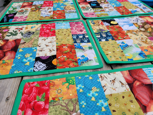 Colorful patchwork quilted placemats with green borders arranged on a wooden surface. The I Spy placemats feature a variety of printed fabric panels including fruit, flowers, insects, and nature scenes. 