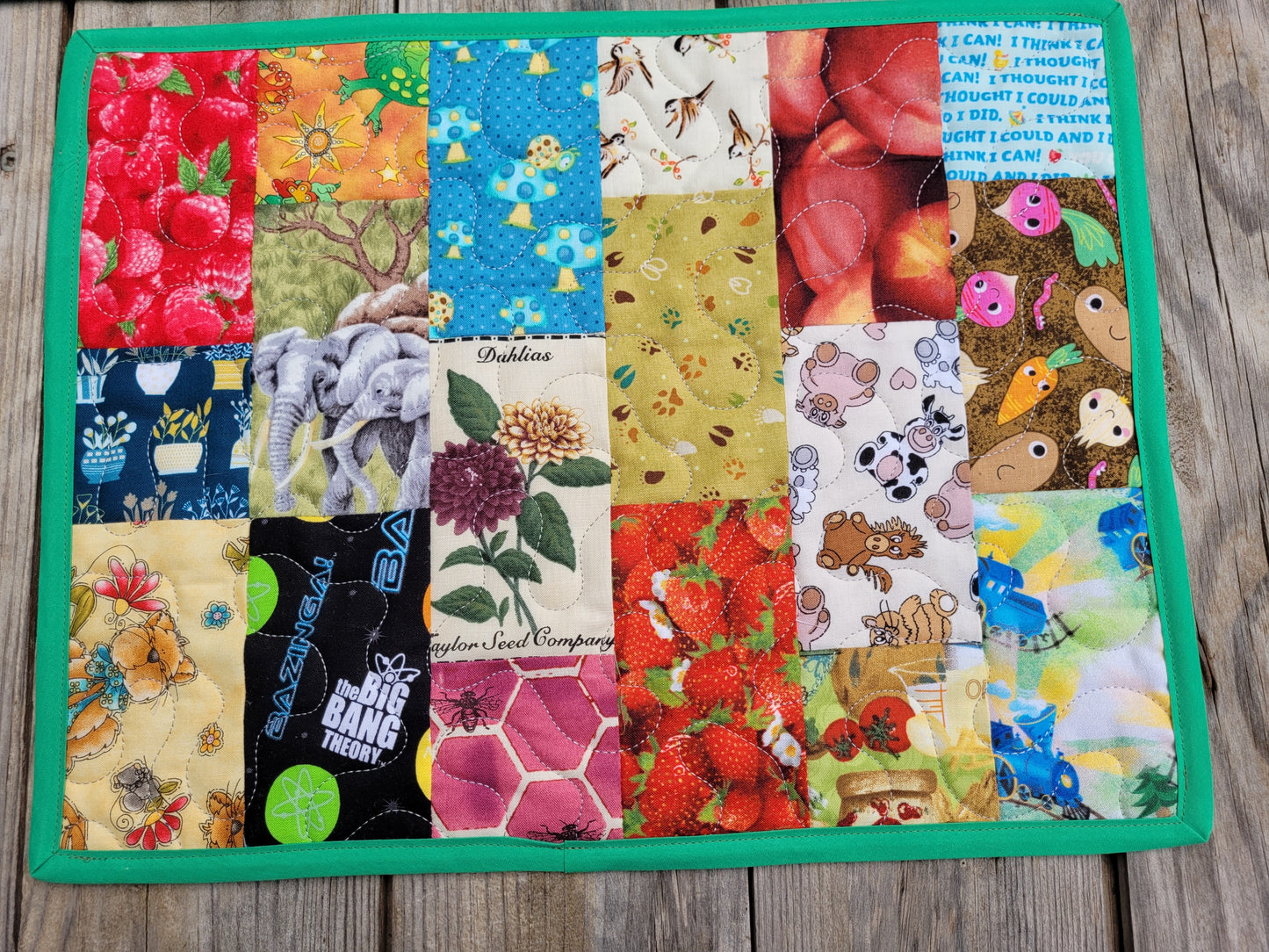 A square quilted fabric placemat with a colorful patchwork design featuring various animal and nature-themed prints, finished with a solid green border, displayed flat on a gray wooden surface.