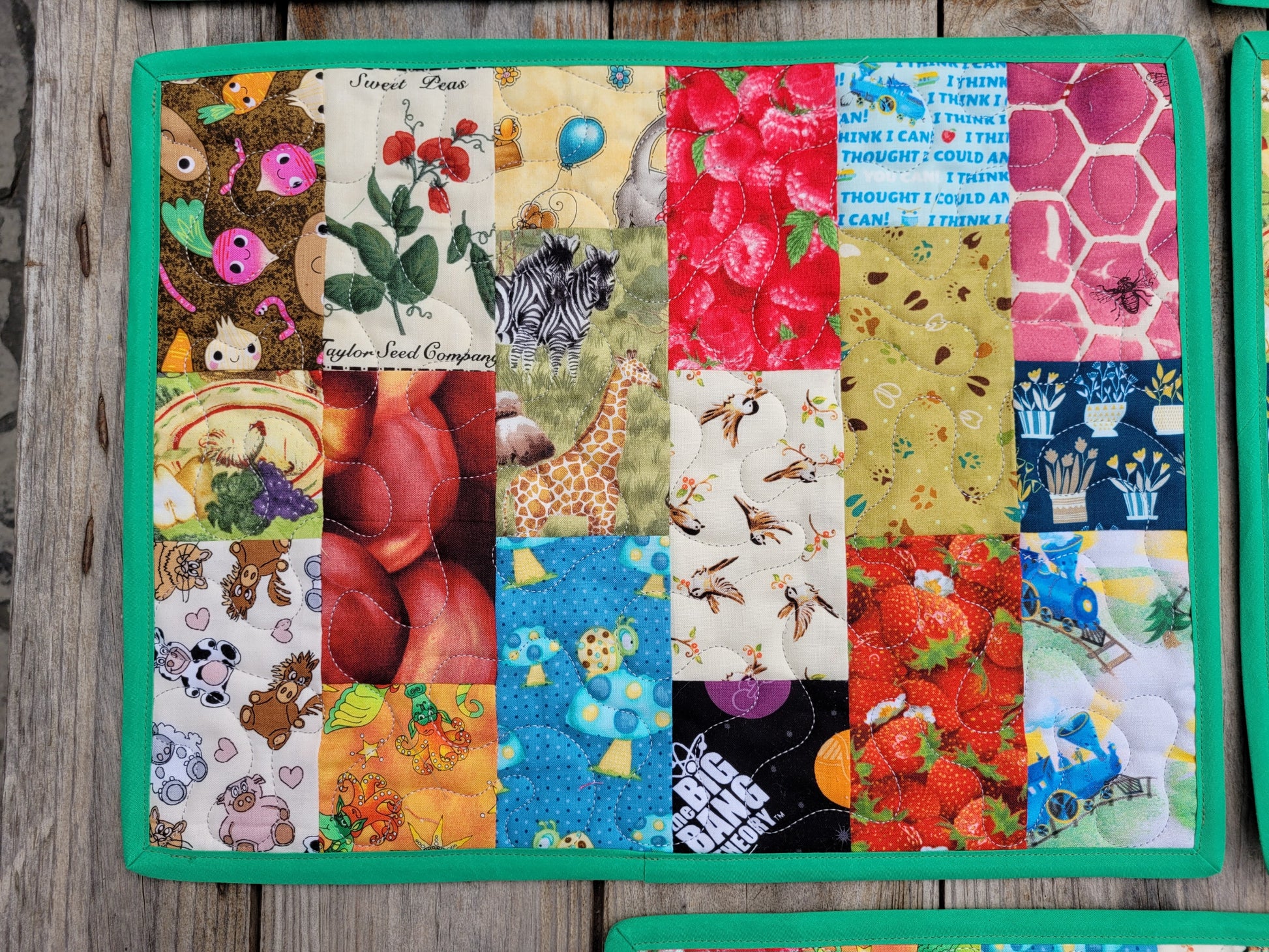 A square quilted fabric placemat, featuring a design of colorful vertical patchwork with various animal, fruit, and nature prints, finished with a solid green border, laid flat on a rustic wooden surface.