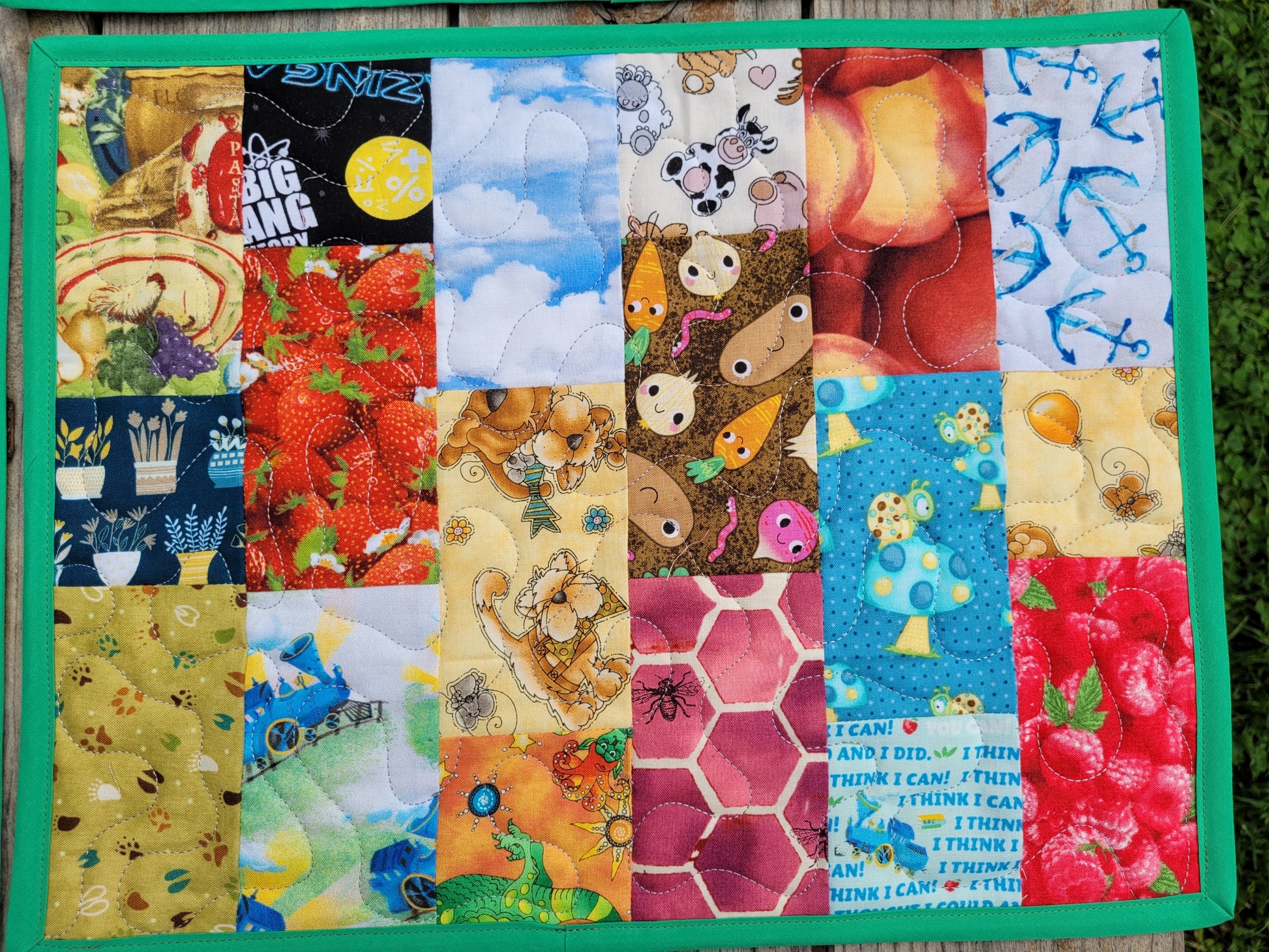 A square quilted patchwork placemat with a bright green border, featuring various colorful cartoon and nature-themed fabrics, photographed outdoors with green grass and a wooden table partially visable in the background.