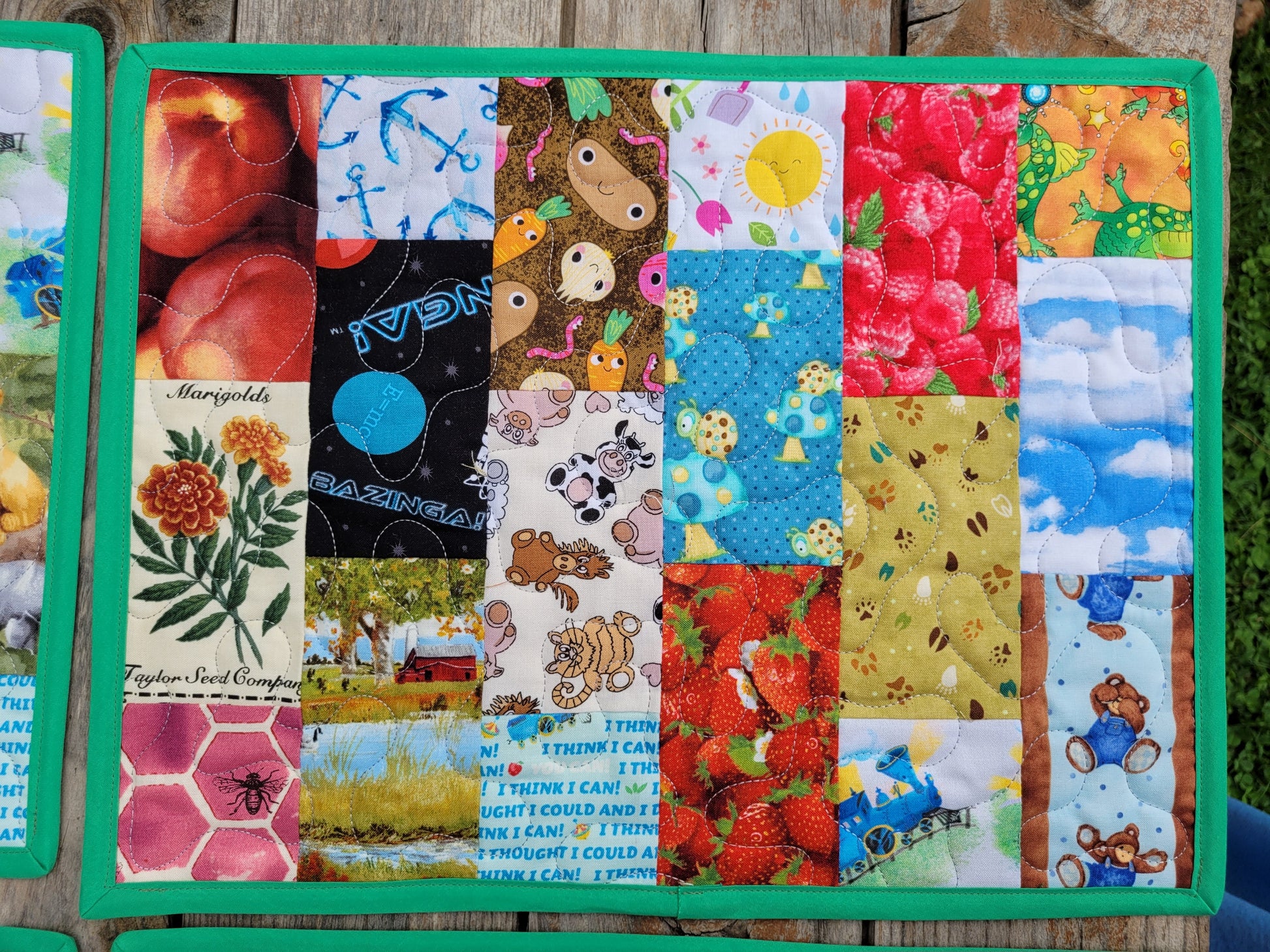 A colorful patchwork quilted fabric placemat, featuring various themed print panels including strawberries, monkeys, space, and flowers, bordered by a bright green fabric edge.