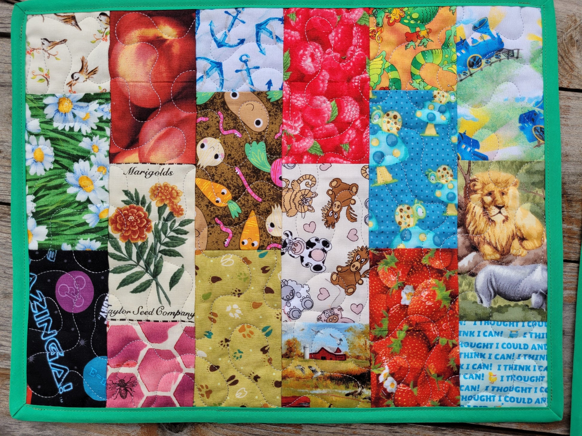 A square quilted fabric mat featuring a colorful patchwork grid of various animal and nature prints, finished with a solid green border, shown on a rustic wooden background.