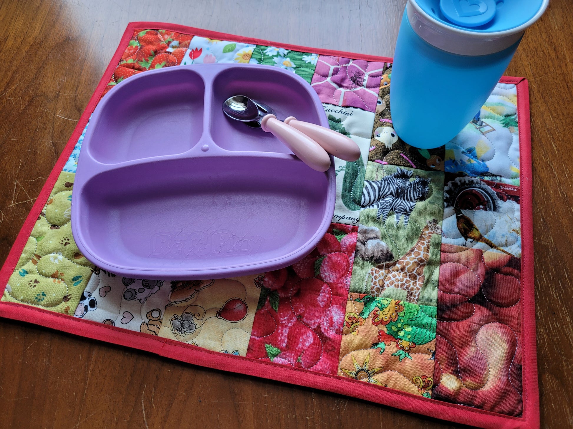 A purple silicone divided baby feeding tray with a pink-handled spoon, placed on a colorful quilted placemat on a wooden table, with a blue tumbler partially visible.