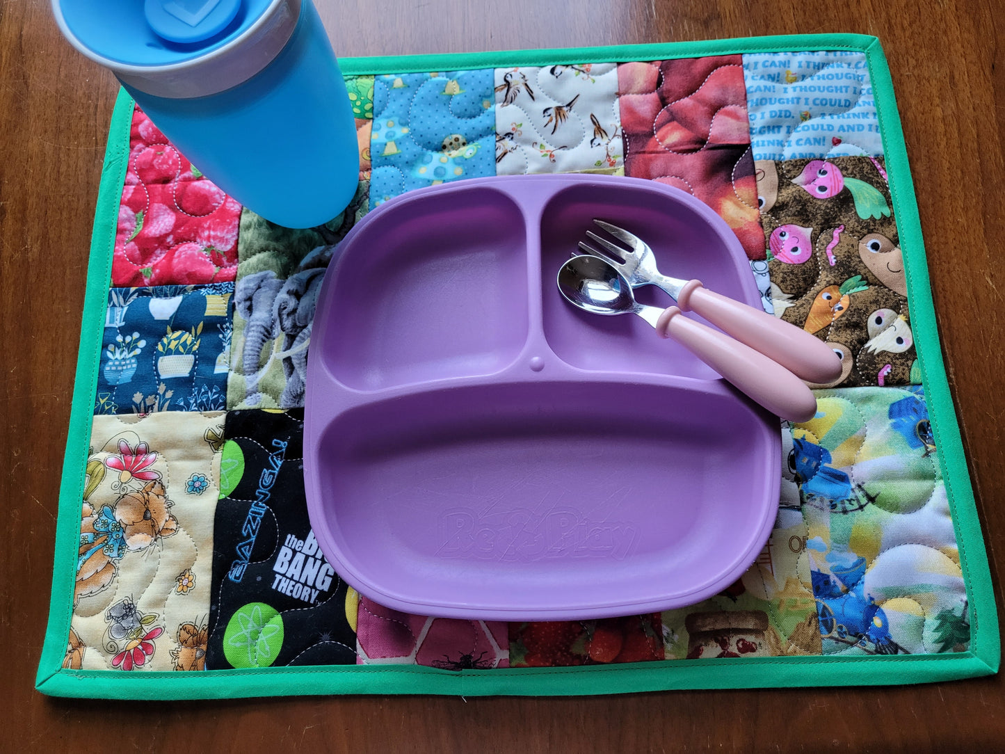 A purple silicone divided baby plate with pink-handled utensils, placed on a colorful patchwork quilt placemat on a wooden table, with a blue cup partially visible in the top left.