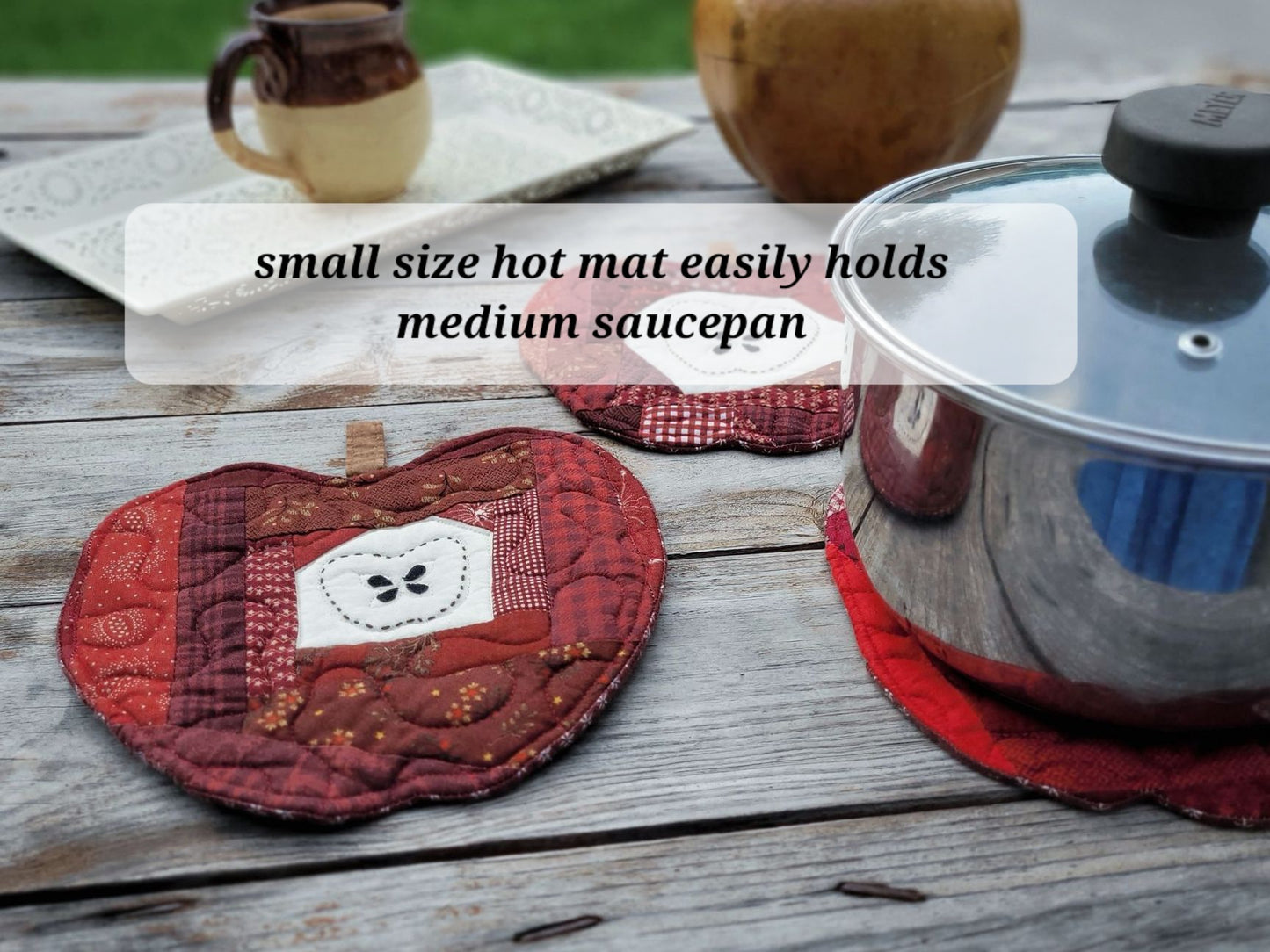 A quilted red and brown apple-shaped hot mat with a white embroidered patch, shown on a rustic wooden table next to a stainless steel saucepan. A text overlay reads 'small size hot mat easily holds medium saucepan'.