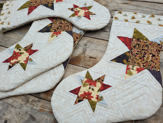 Four quilted Christmas stockings with star patterns, featuring a mix of cream, red, green, and brown fabrics, arranged on a rustic wooden surface.