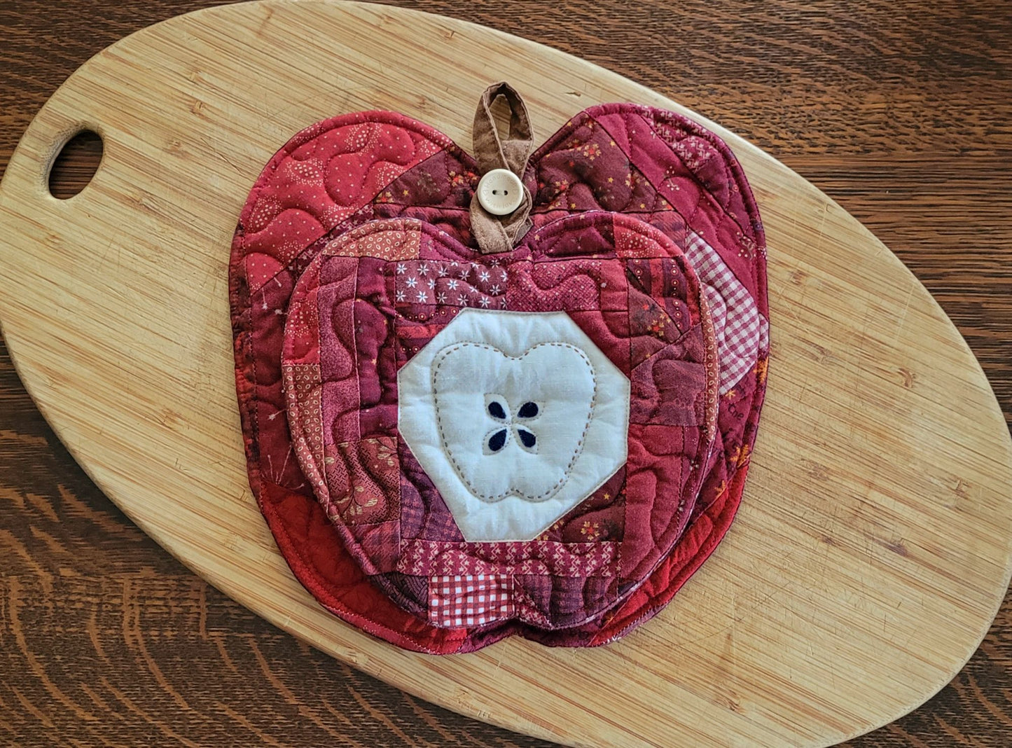Two quilted fabric apple-shaped mini quilts  in shades of red and burgundy, featuring a white fabric apple core with black stitched seeds displayed on a light wood cutting board over a dark wood table. The apples are nested on top of each other using the button hanging loop on the larger apple to hang the smaller one in front as an appealing gift set. 