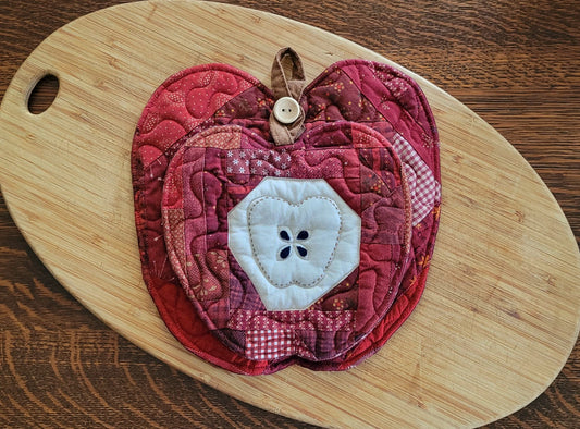Two quilted fabric apple-shaped mini quilts  in shades of red and burgundy, featuring a white fabric apple core with black stitched seeds displayed on a light wood cutting board over a dark wood table. The apples are nested on top of each other using the button hanging loop on the larger apple to hang the smaller one in front as an appealing gift set. 
