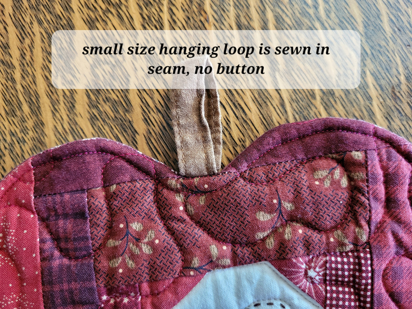 Close-up of a quilted fabric apple-shaped quilt in red, burgundy, and floral patchwork fabrics, with a sewn-in loop at the top and a text overlay reading 'small size hanging loop is sewn in, no button'.