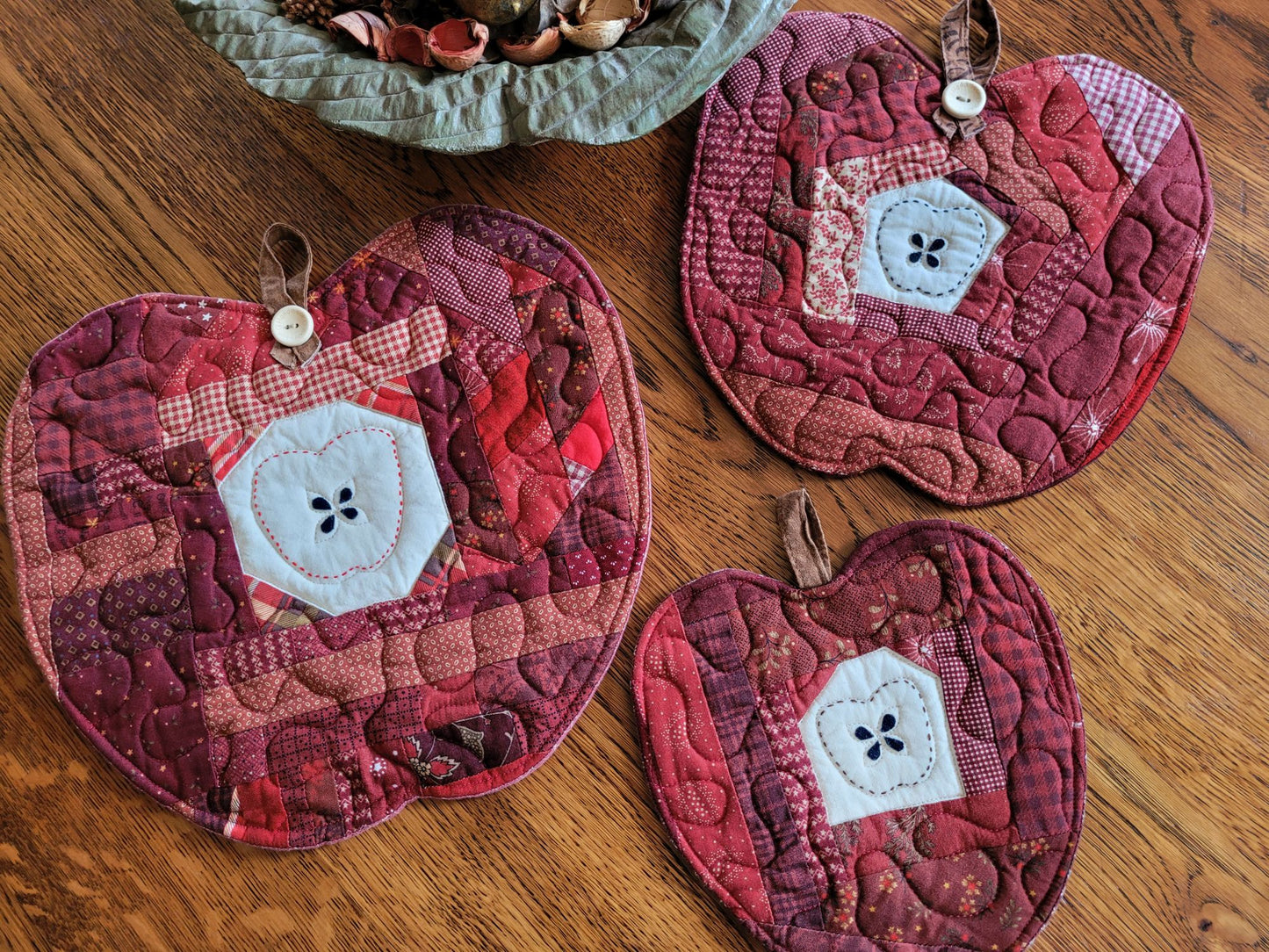 Three quilted apple-shaped mats in shades of red and burgundy are arranged on a wooden table. Each mat features a centre motif with a hand-stitched apple  outline around applique seeds on a light muslin fabric, a brown hanging loop is attached at the top with a wooden button stamped with 'handmade with love'.
