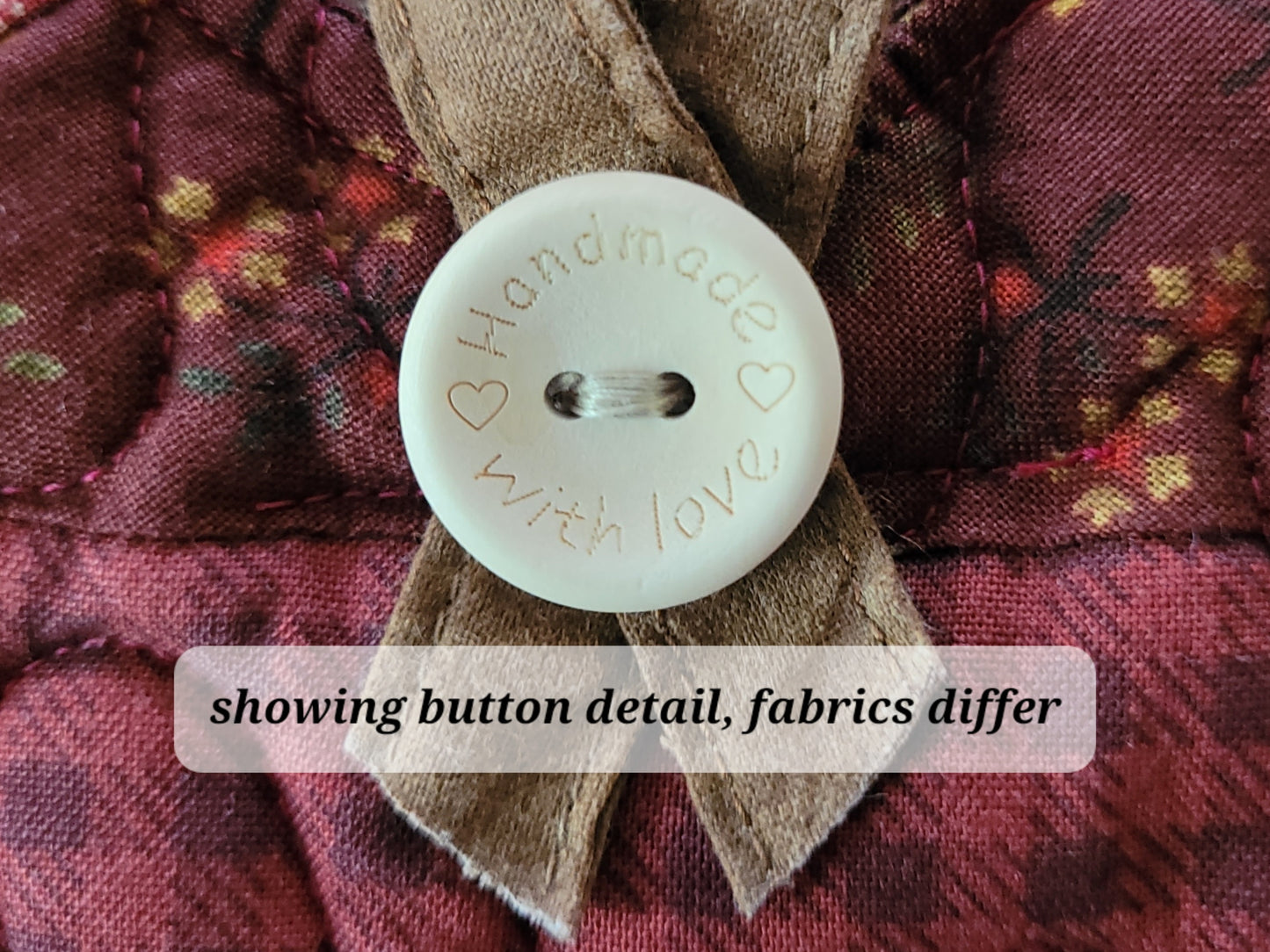 Close-up of a round off-white wooden button with 'Handmade with love' stamped on it, set against a quilted burgundy fabric with a brown hanging loop. A text overlay at the bottom reads 'showing button detail, fabrics differ'.