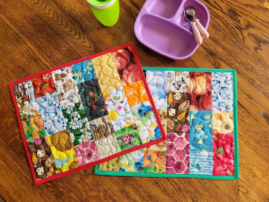 Two Quilted I Spy Placemats for kids on a wooden table with a child's purple divided plate with pink cutlery and a green sippy cup partially visable.