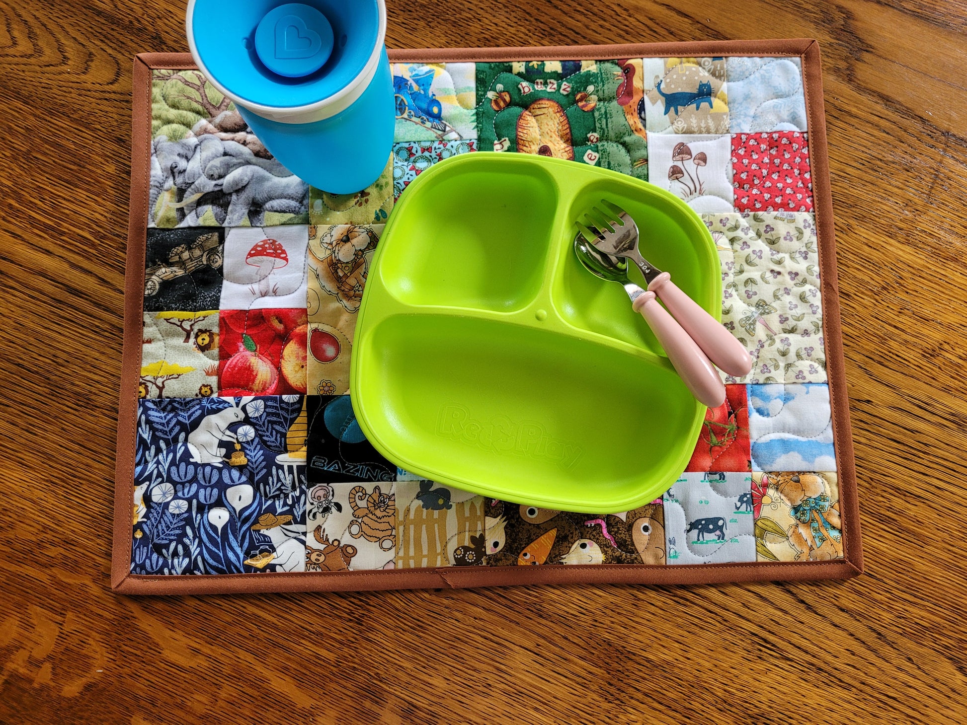 A bright lime green three-compartment divided baby plate with pink-handled utensils, arranged on a colorful patchwork placemat over a wooden table. A blue cup is partially visible at the top left.