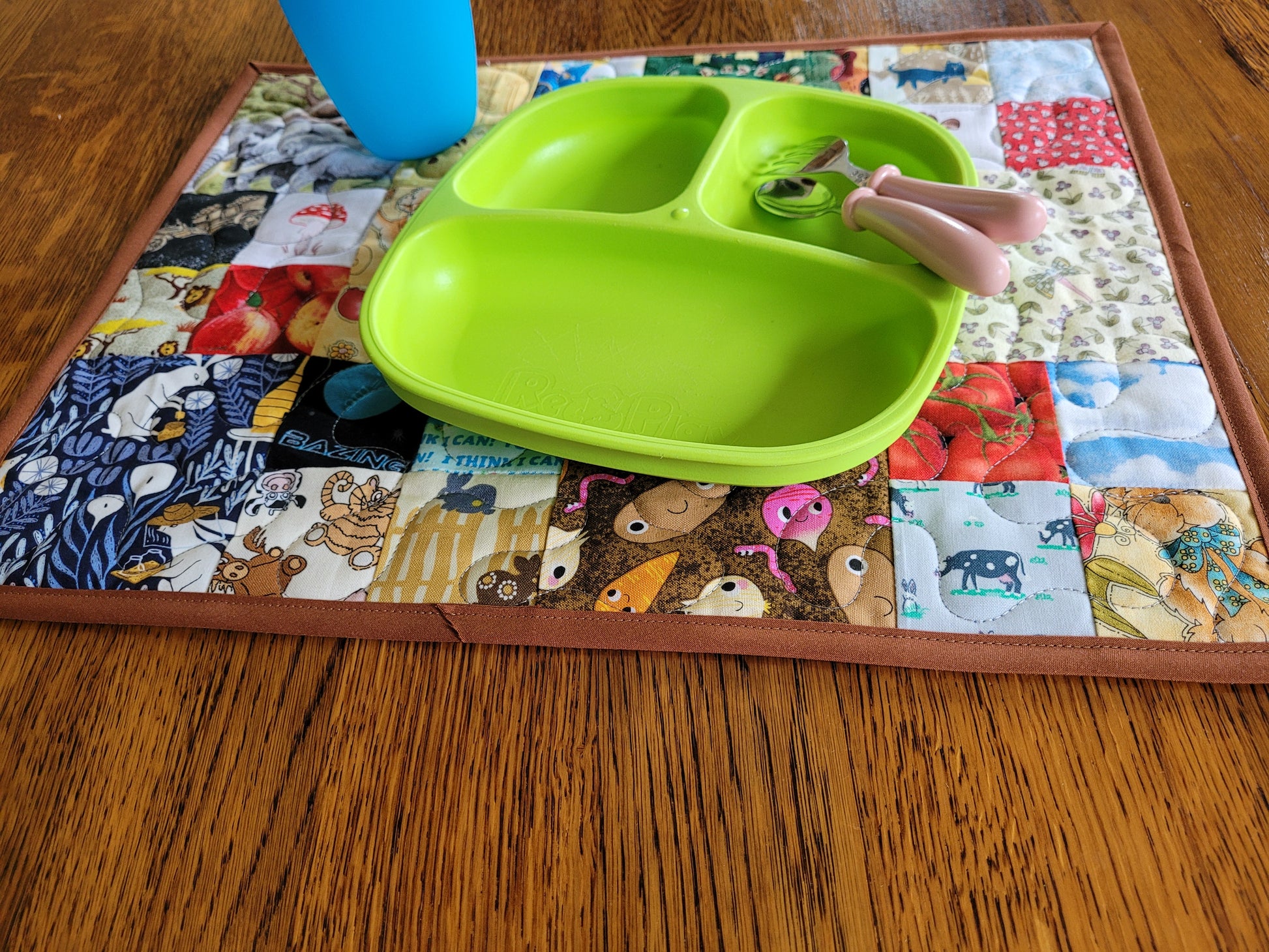 A bright lime green divided baby plate with two pink-handled utensils, placed on a colorful patchwork quilt placemat on a wooden table. A blue cup is partially visible at the top.