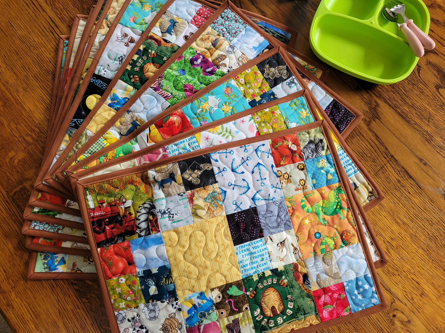 A fan of colorful quilted placemats with brown borders arranged on a wooden table, next to a bright green divided baby feeding tray with a spoon.