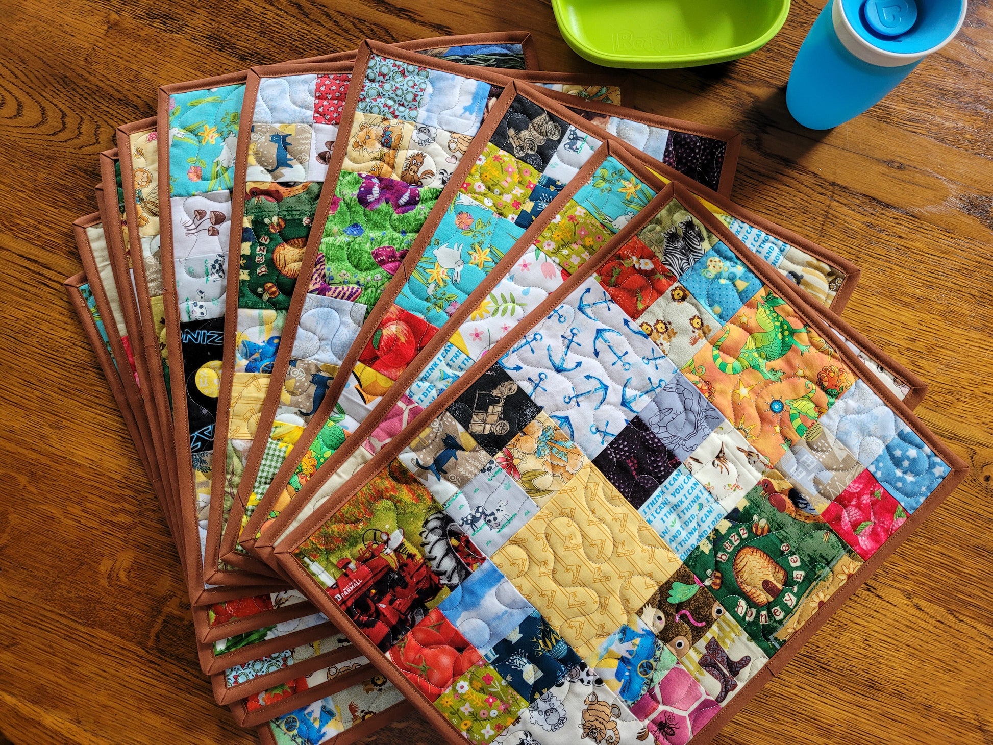 A stack of colorful quilted placemats with brown borders, fanned out on a wooden table next to a green plate and a blue cup.