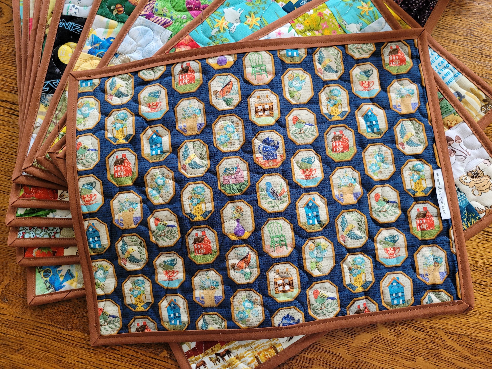 Showing the back of a quilted placemat in a navy blue background fabric with a repeating pattern of small illustrated scenes inside octagonal frames, surrounded by a brown binding. Multiple similar placemats are fanned out on a wooden table.