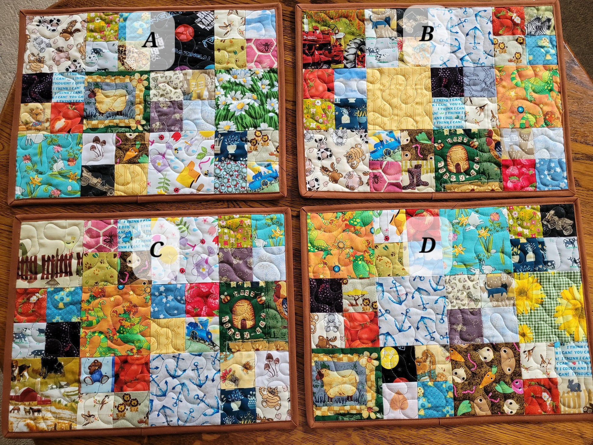 Four square quilted patchwork placemats laid out on a wooden surface, labeled A, B, C, and D. Each piece features a colorful patchwork design with various animal, floral, and nature-themed fabrics.