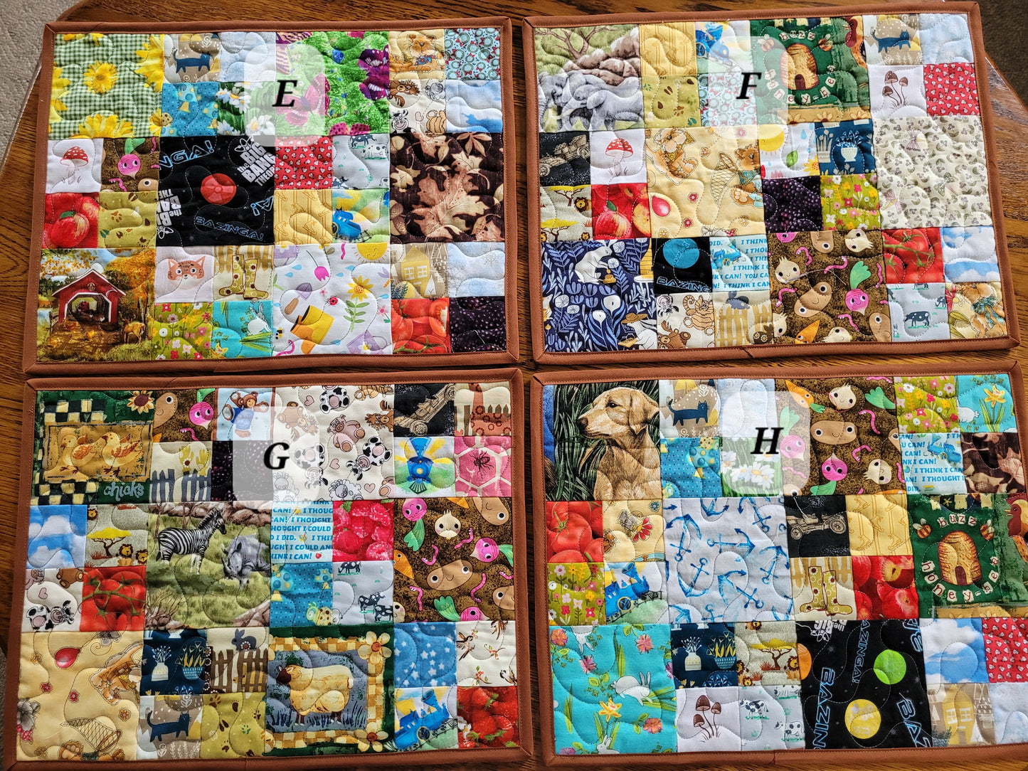 Four handmade quilted placemats labeled E, F, G, and H arranged in a 2x2 grid. Each mat features a colorful patchwork design with various baby-themed prints including teddy bears, animals, fruits, and nature scenes, all surrounded by a brown binding.