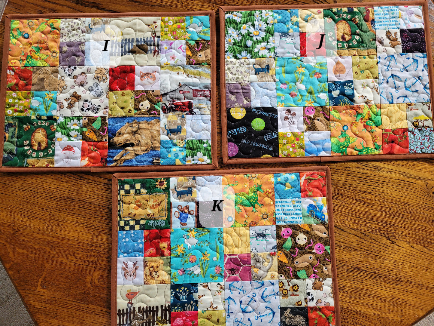 Three I SPy patchwork placemats labeled I, J, and K displayed on a wooden table. Each mat features a colorful grid of various fabric patches with whimsical prints, including animals, nature scenes, and patterns.
