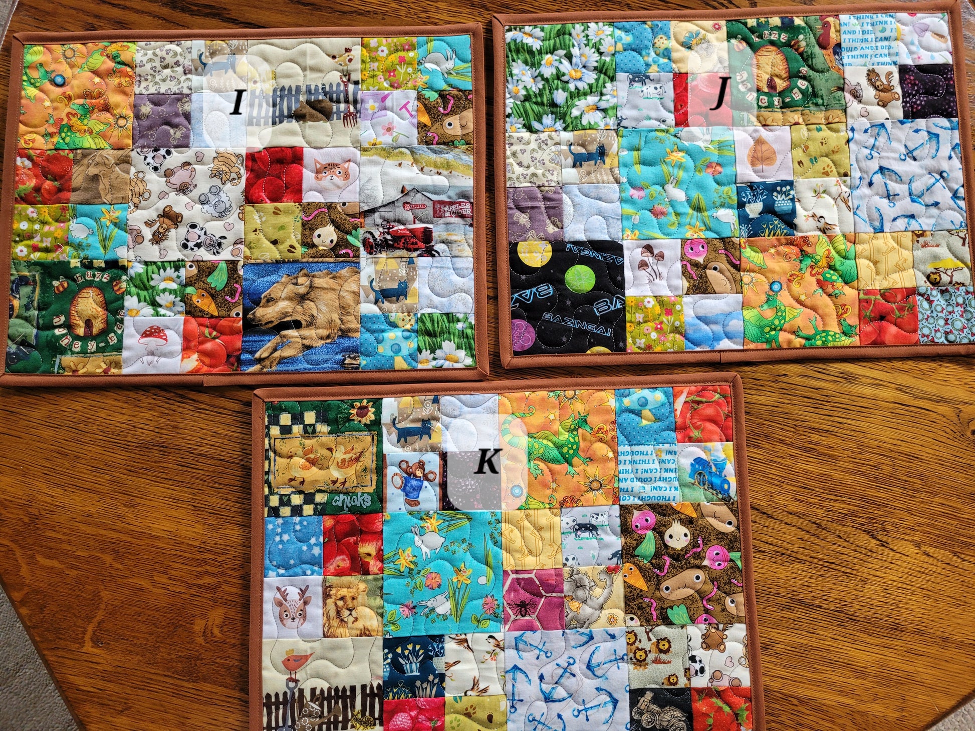 Three I SPy patchwork placemats labeled I, J, and K displayed on a wooden table. Each mat features a colorful grid of various fabric patches with whimsical prints, including animals, nature scenes, and patterns.