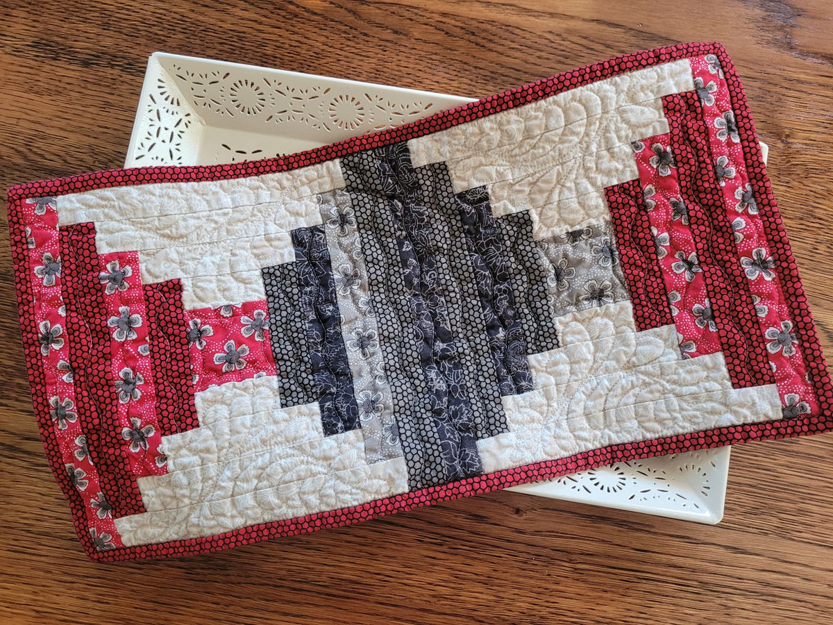 A rectangular quilted table topper with a geometric log cabin pattern, featuring red and gray fabric panels on a white quilted background, displayed on a white decorative tray over a wooden table.