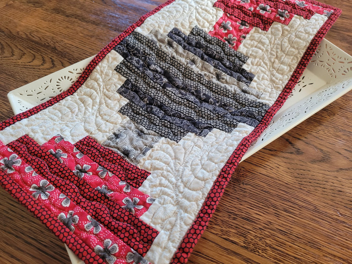 A handmade quilted table runner with a rustic log cabin style design, featuring red and white floral patterned sections, dark gray patterned stripes, and white quilted sections with red borders, displayed on a white decorative tray over a wooden table.