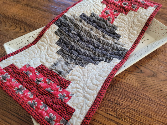 A handmade quilted table runner with a rustic log cabin style design, featuring red and white floral patterned sections, dark gray patterned stripes, and white quilted sections with red borders, displayed on a white decorative tray over a wooden table.