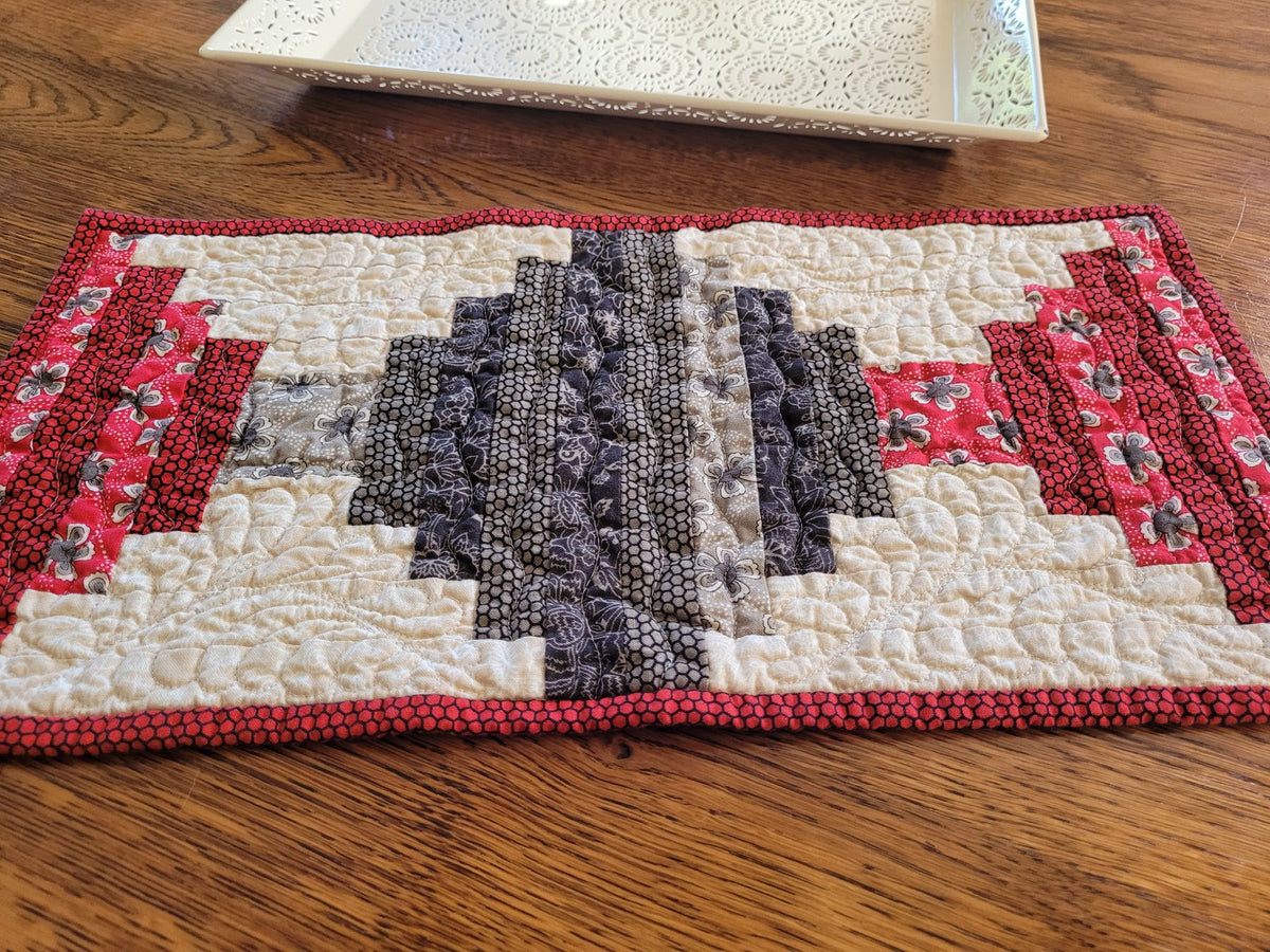 Small quilted table runner in black, gray and red log cabin style patchwork on a wooden table with a white decorative tray in the background.