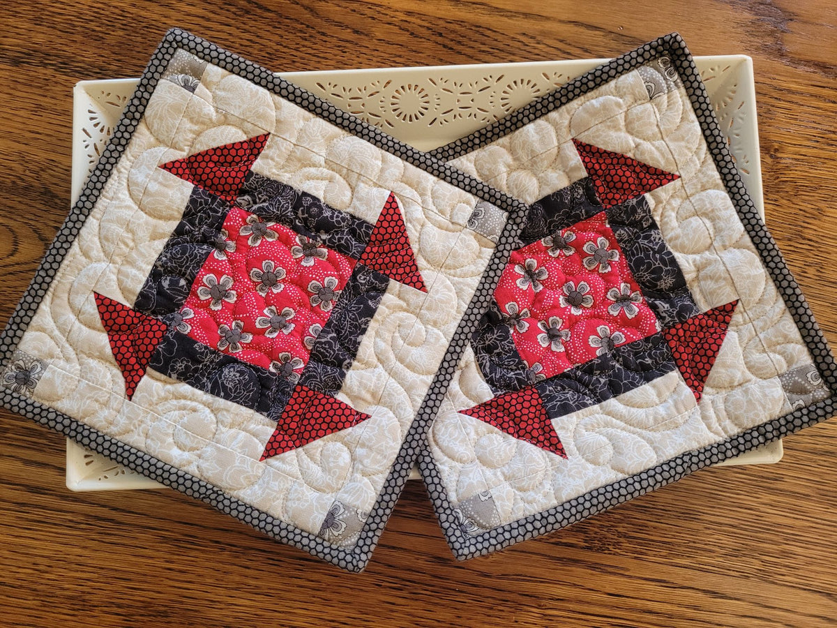 Two quilted fabric potholders with a red, black, and beige block pattern, displayed on a cream-colored tray over a wooden table.