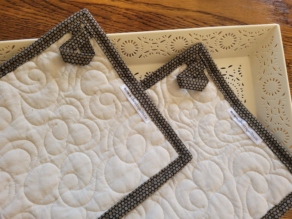 Two quilted fabric potholders showing the back with hanging loops and brand labels, displayed on a cream-colored tray over a wooden table.