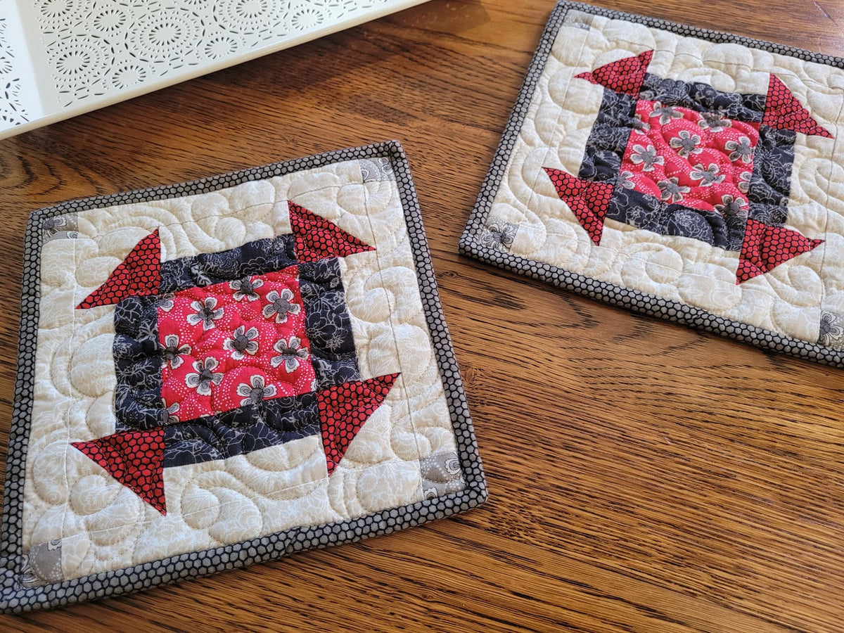 Two handmade quilted fabric potholders with a geometric block pattern, displayed on a wooden tabletop.