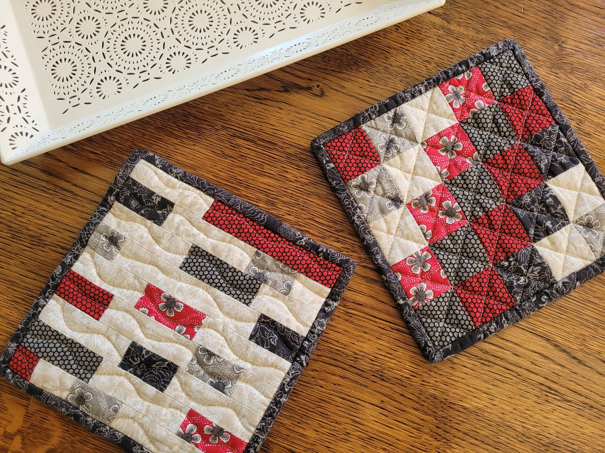 Two handmade quilted fabric potholders with red, black, and cream patchwork designs, displayed on a wooden tabletop next to a white decorative tray.