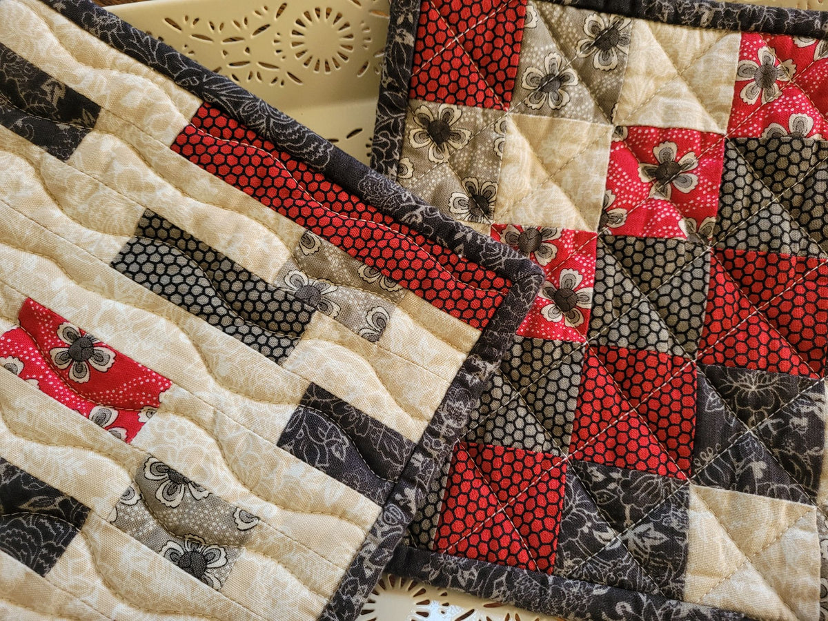 A close-up of quilted patchwork hot mats with cream, red, black, and taupe fabric squares, including floral and geometric patterns, laid over a light-colored surface.