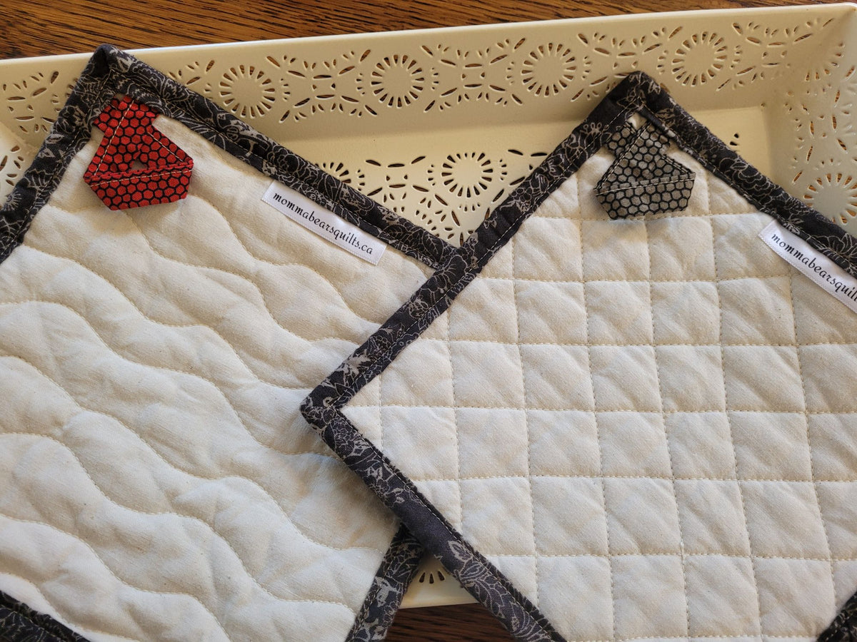 Two quilted fabric potholders showing the back side with hanging loops and brand labels, displayed on a cream-colored tray.