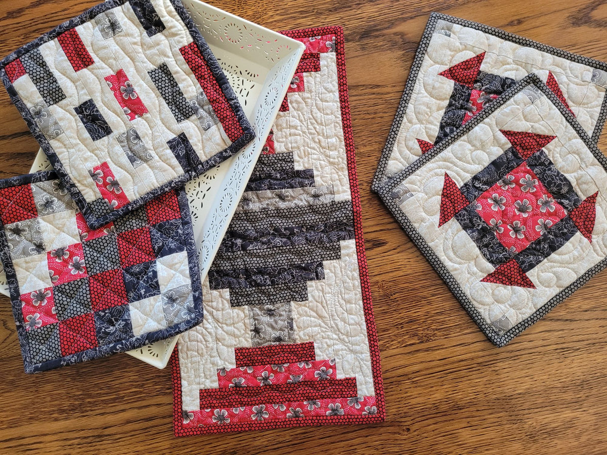 A coordinating quilted table set including a small table runner and two sets of kitchen hot mats styled on a wooden table with a white decorative tray . 