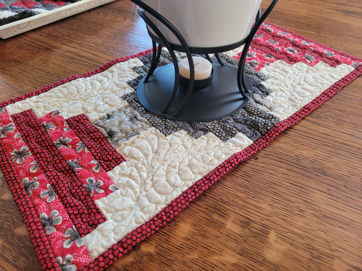A rectangular quilted table runner with a rustic log cabin pattern in red, cream, and gray, styled on a wooden table with a black metal candle holder on top.