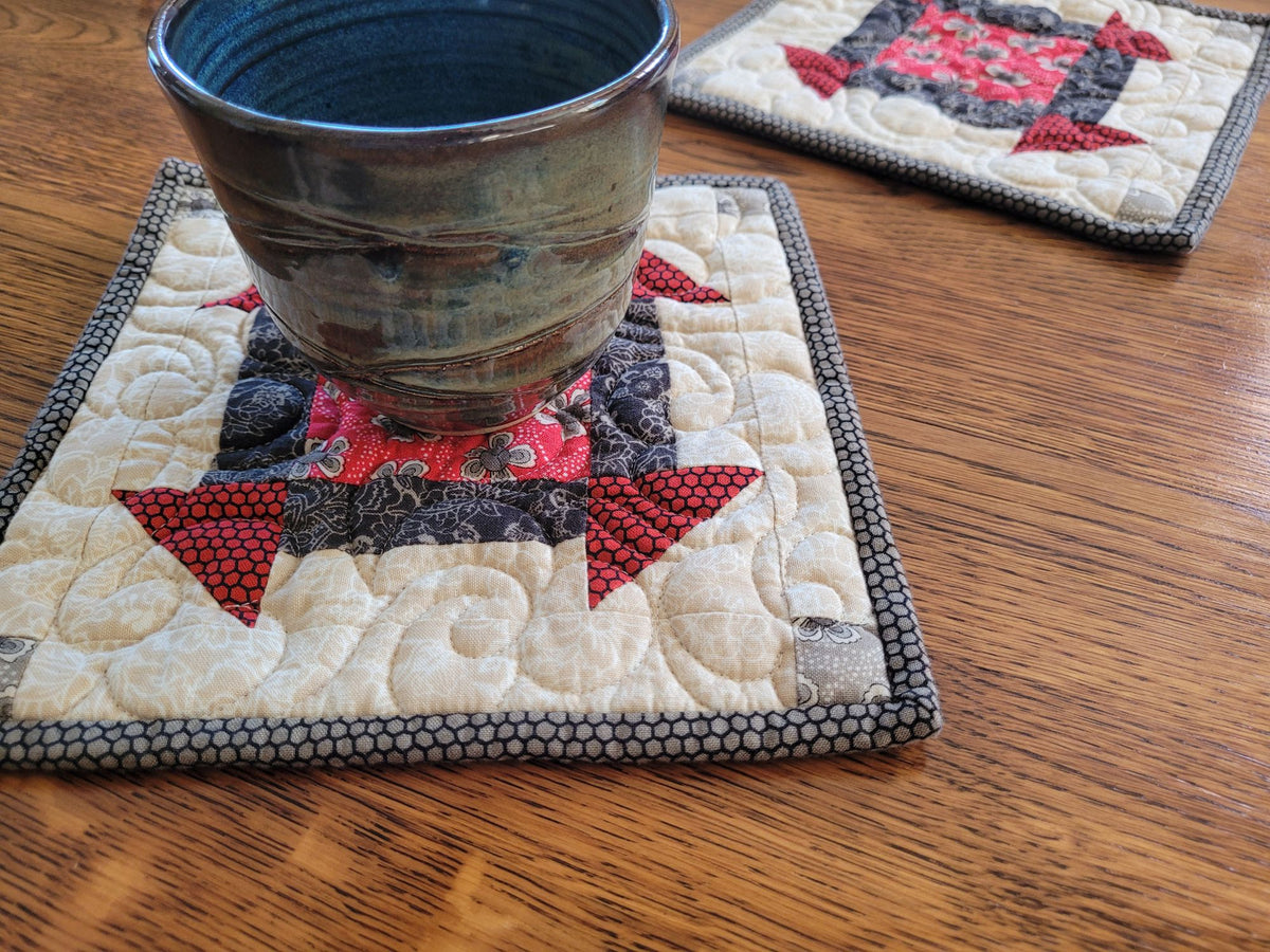 A close-up of a handmade quilted patchwork potholder, holding a dark glazed ceramic mug on a wooden table. A second matching potholder is visible in the background.
