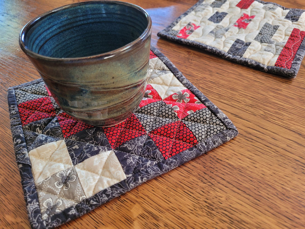 A close-up of a handmade quilted hot mat in red, gray, and cream, holding a dark glazed ceramic mug on a wooden table. A second matching hot mat is visible in the background.