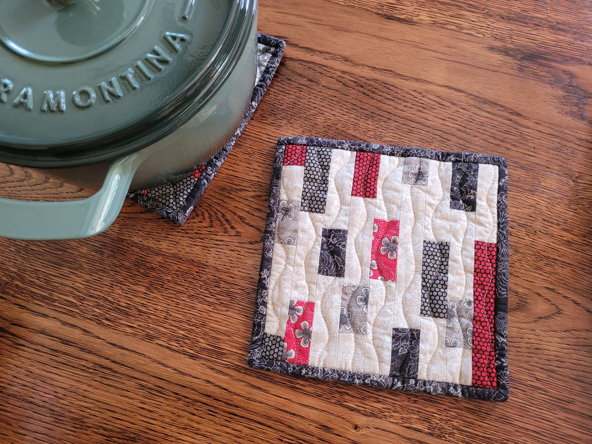 A square quilted fabric trivet or hot mat with a patchwork design in cream, red, black, and gray, shown on a wooden table next to a green cast iron pot.