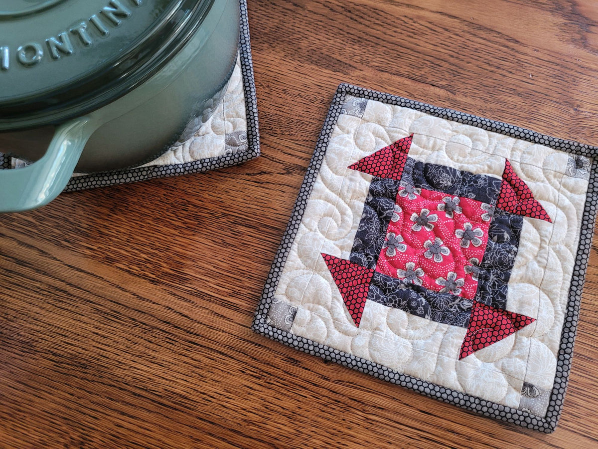 A square quilted fabric trivet or potholder with a geometric block pattern in red, black, and cream, shown on a wooden table next to a teal pot.
