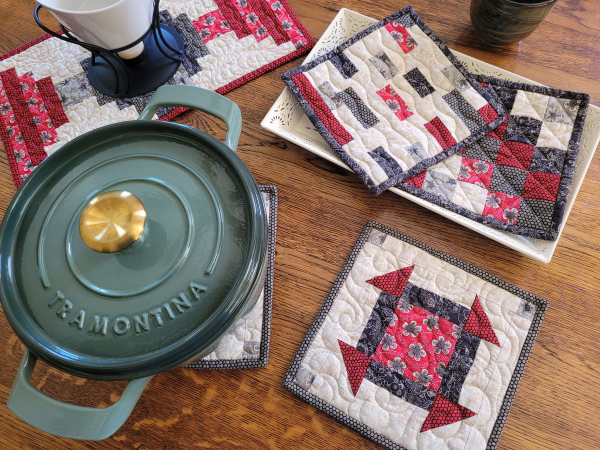 A set of handmade quilted cotton table linens, including two sets of hot mats and a small runner, displayed on a wooden table alongside a green enameled cast iron pot and ceramic dishes.