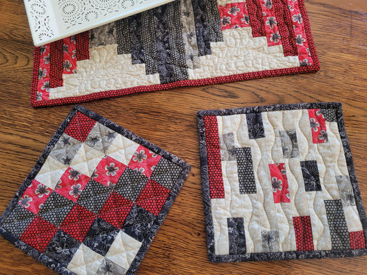 A quilted table set including a small table runner with two coordinating hot mats on a wooden table with a white decorative tray partially visable. 