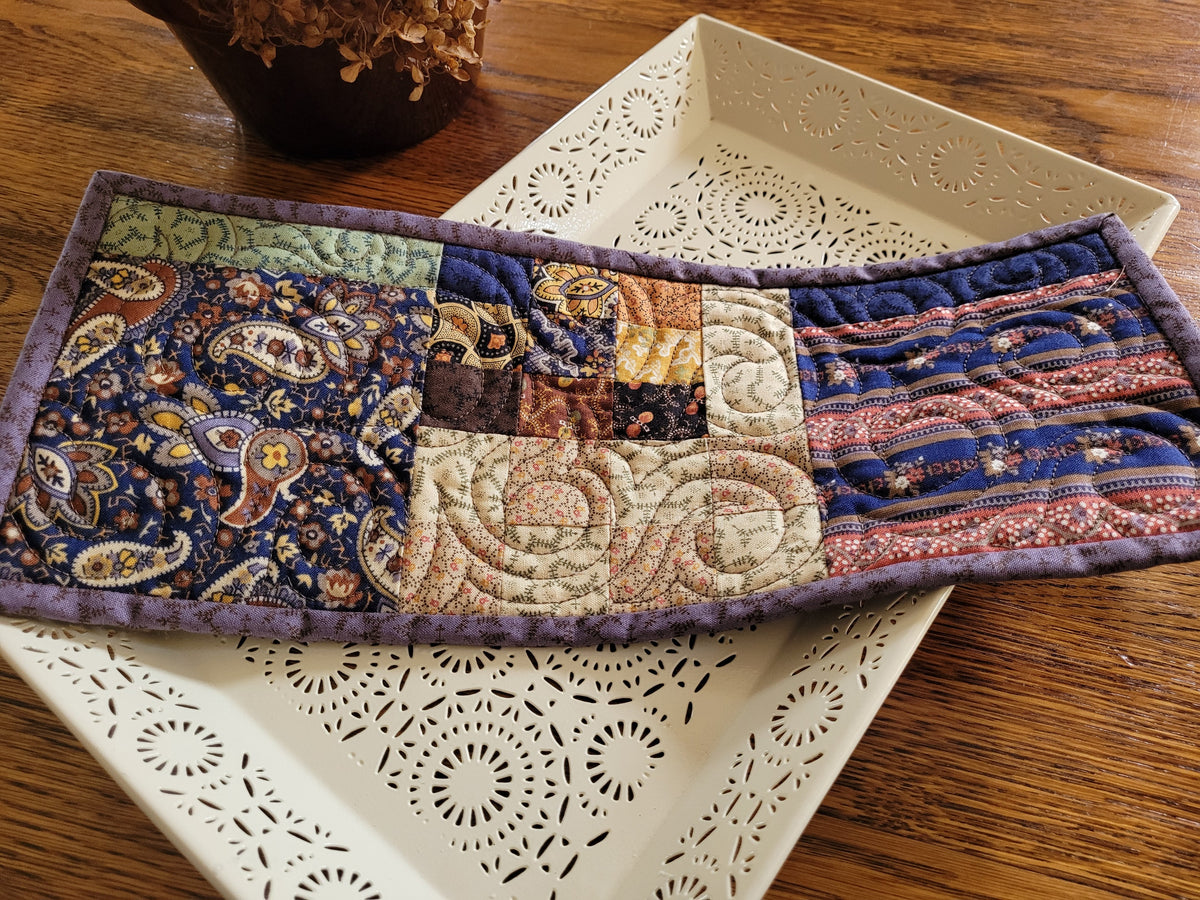 A small rectangular patchwork quilt with a purple border, laid diagonally across a white decorative tray on a wooden table.
