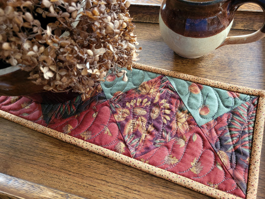 A small rectangular quilt with a rustic tan border, featuring a patchwork design of red, burnt orange and green leaf patterns, styled on a wooden tray next to a ceramic mug and dried flowers.
